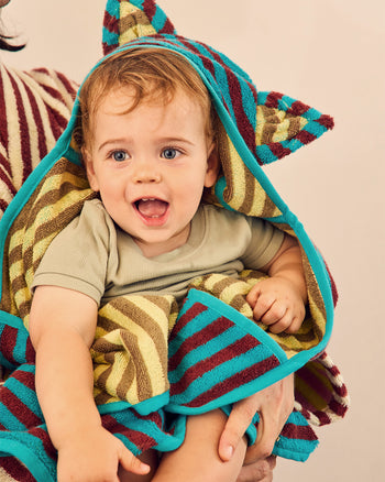 A baby wrapped in a colorful striped hooded towel with ears, held by an adult against a plain beige background. 