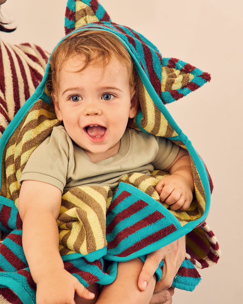 A baby wrapped in a colorful striped hooded towel with ears, held by an adult against a plain beige background. 