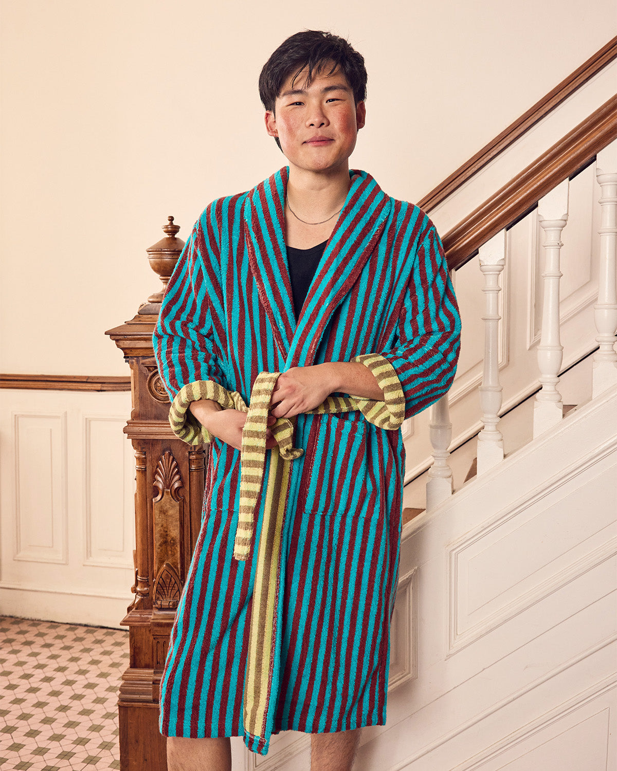 A man wearing a vibrant blue and maroon vertically striped bathrobe with gold trim, standing in front of an indoor staircase.