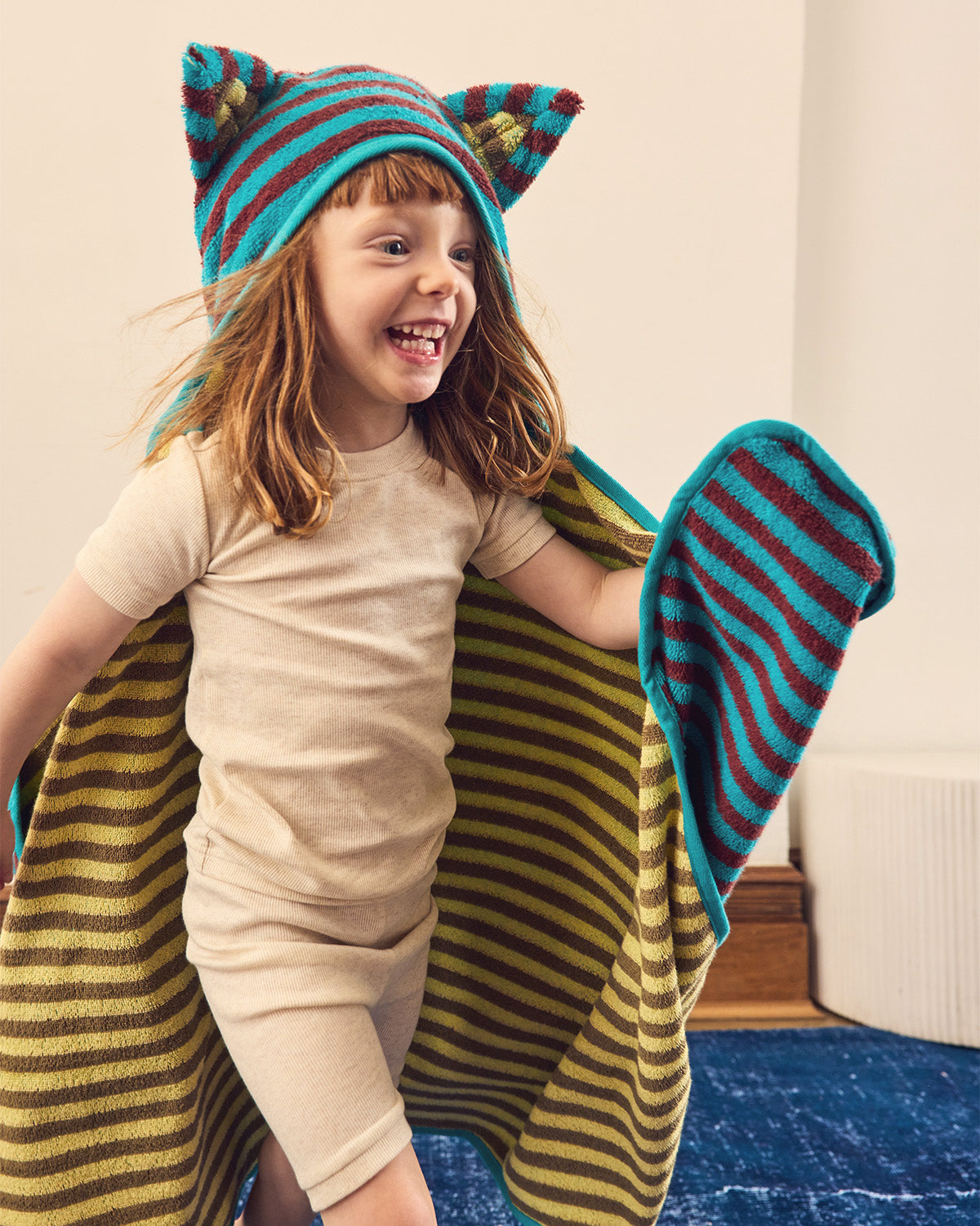 A young child wearing a beige short-sleeve romper and a colorful striped hooded towel hat with  ears, while holding up a matching striped bath towel in a home setting.