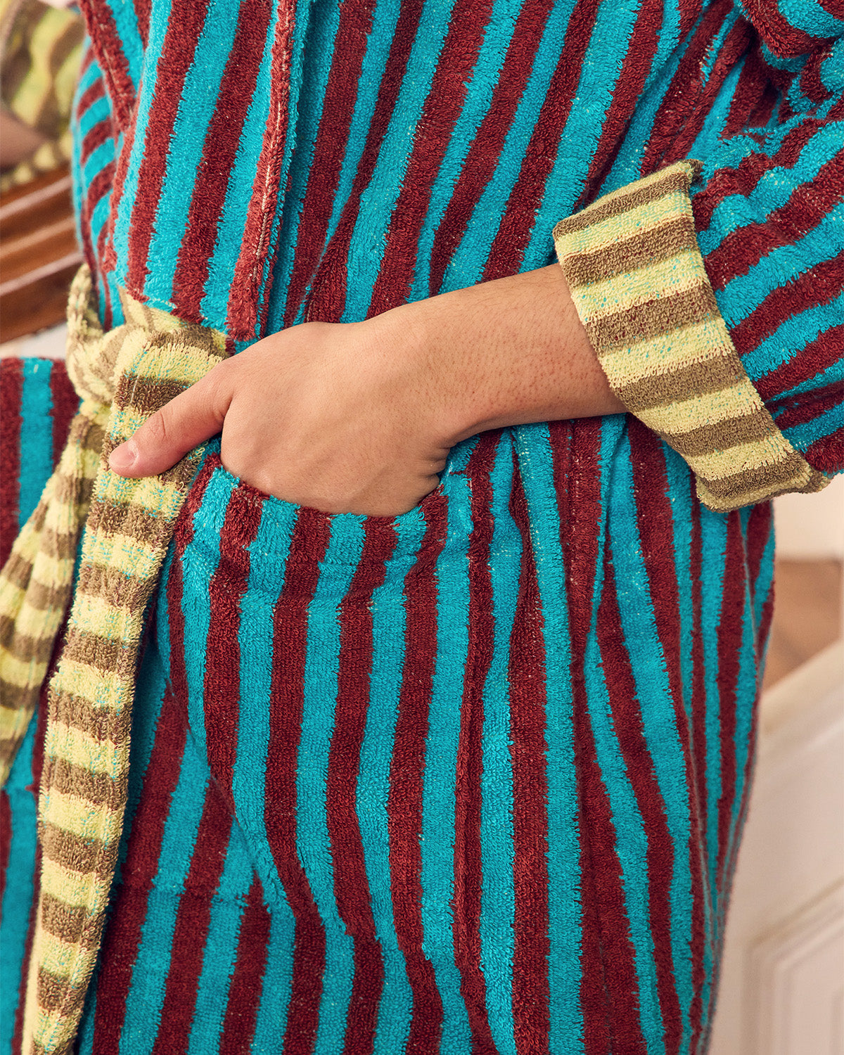 Close-up of a striped bathrobe with a person's hand in the pocket, featuring turquoise, burgundy, and tan vertical stripes with a matching tie belt.