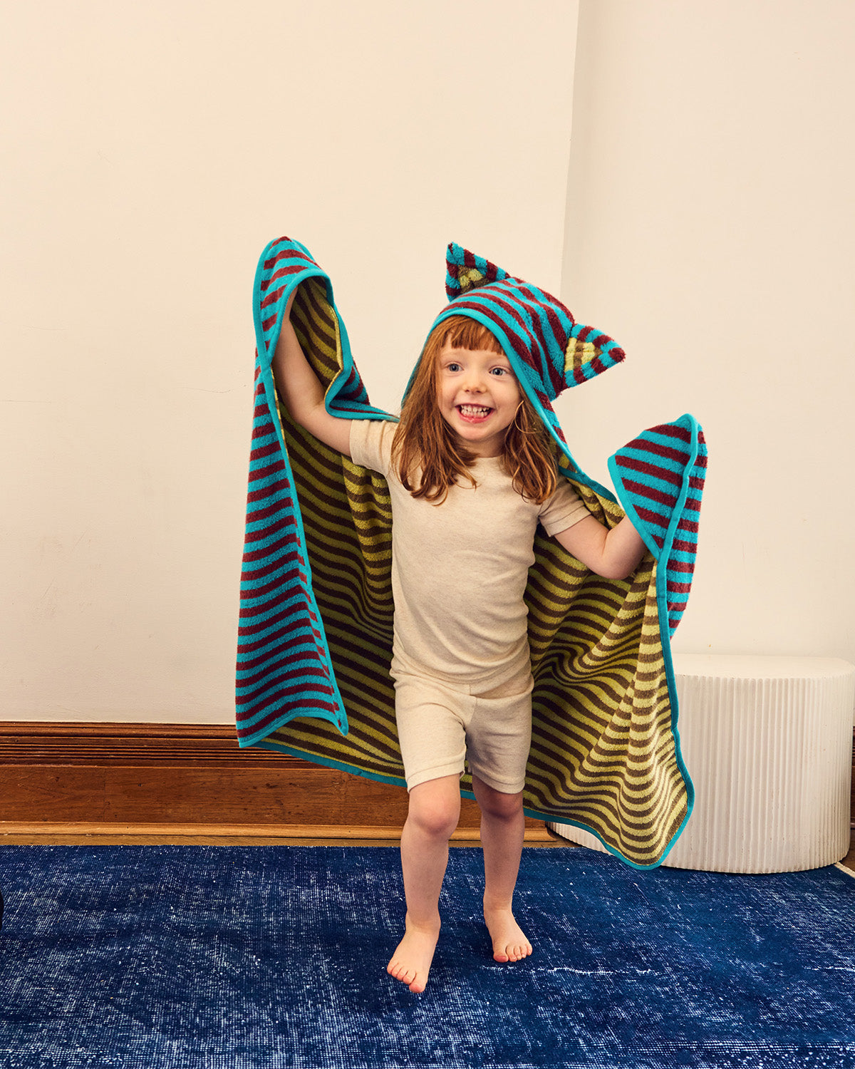 A young child stands barefoot indoors, holding up a hooded towel with striped teal, maroon, beige, and sand. The child is wearing a light beige t-shirt and shorts, set against a plain cream wall and blue rug.