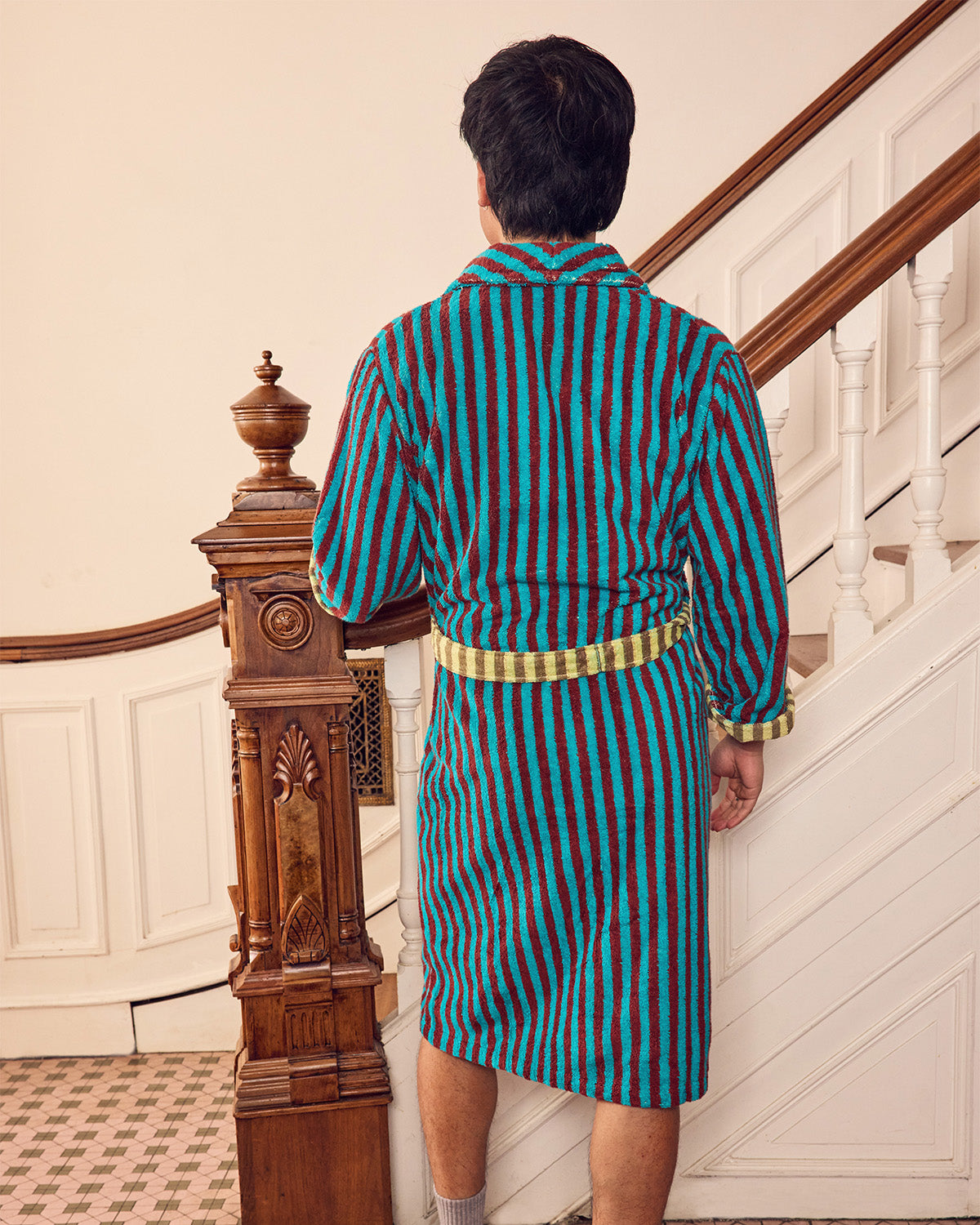 A man viewed from behind wearing a striped bathrobe with teal, red, and black vertical stripes and a yellow belt, standing in front of an indoor staircase.