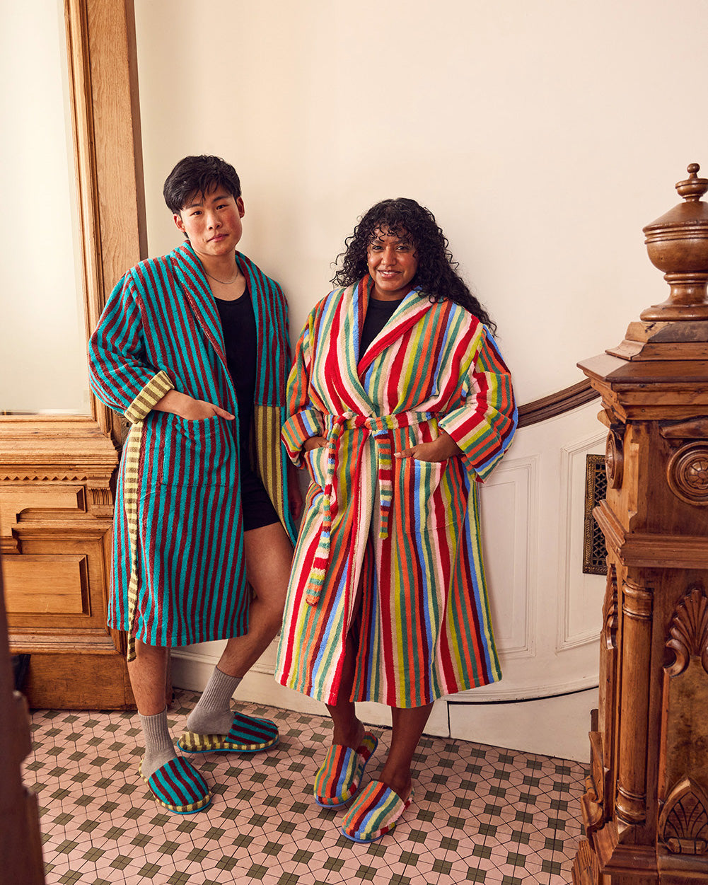 Two models wearing colorful vertically striped bathrobes and matching slippers, standing in an indoor room with a tiled floor and wooden furniture.