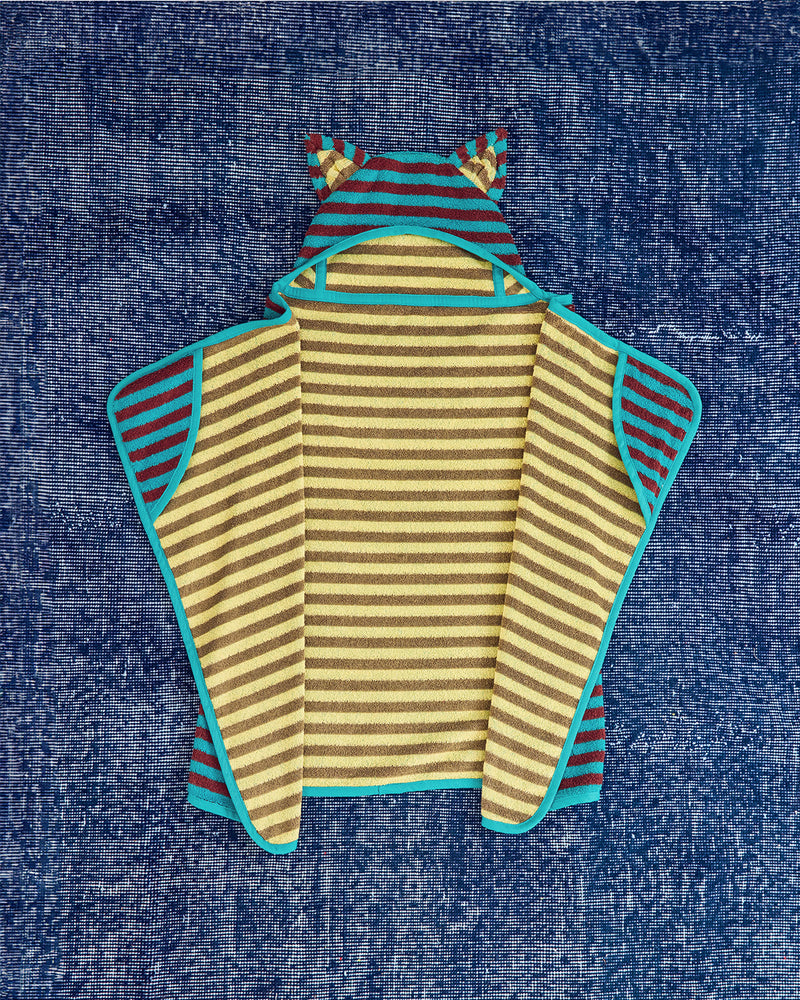 A baby hooded cape  laid flat on a textured blue fabric background. The garment features horizontal yellow and brown stripes on the main body, teal trim, and sand and brown stripes on the hood and side panels.