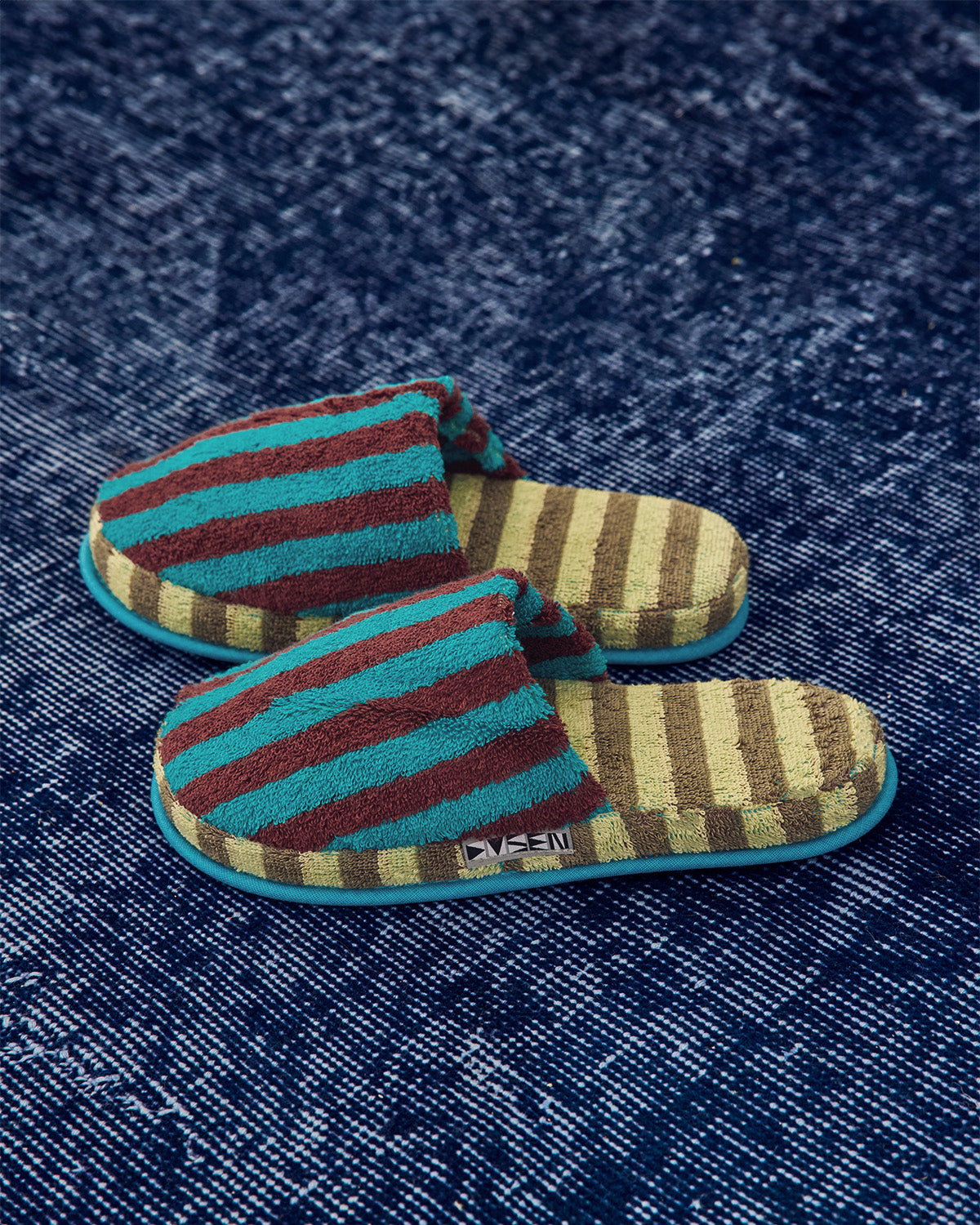 A pair of striped slip-on slippers with a textured terry cloth upper, featuring teal, dark red, and beige horizontal stripes, resting on a dark blue textured rug. 