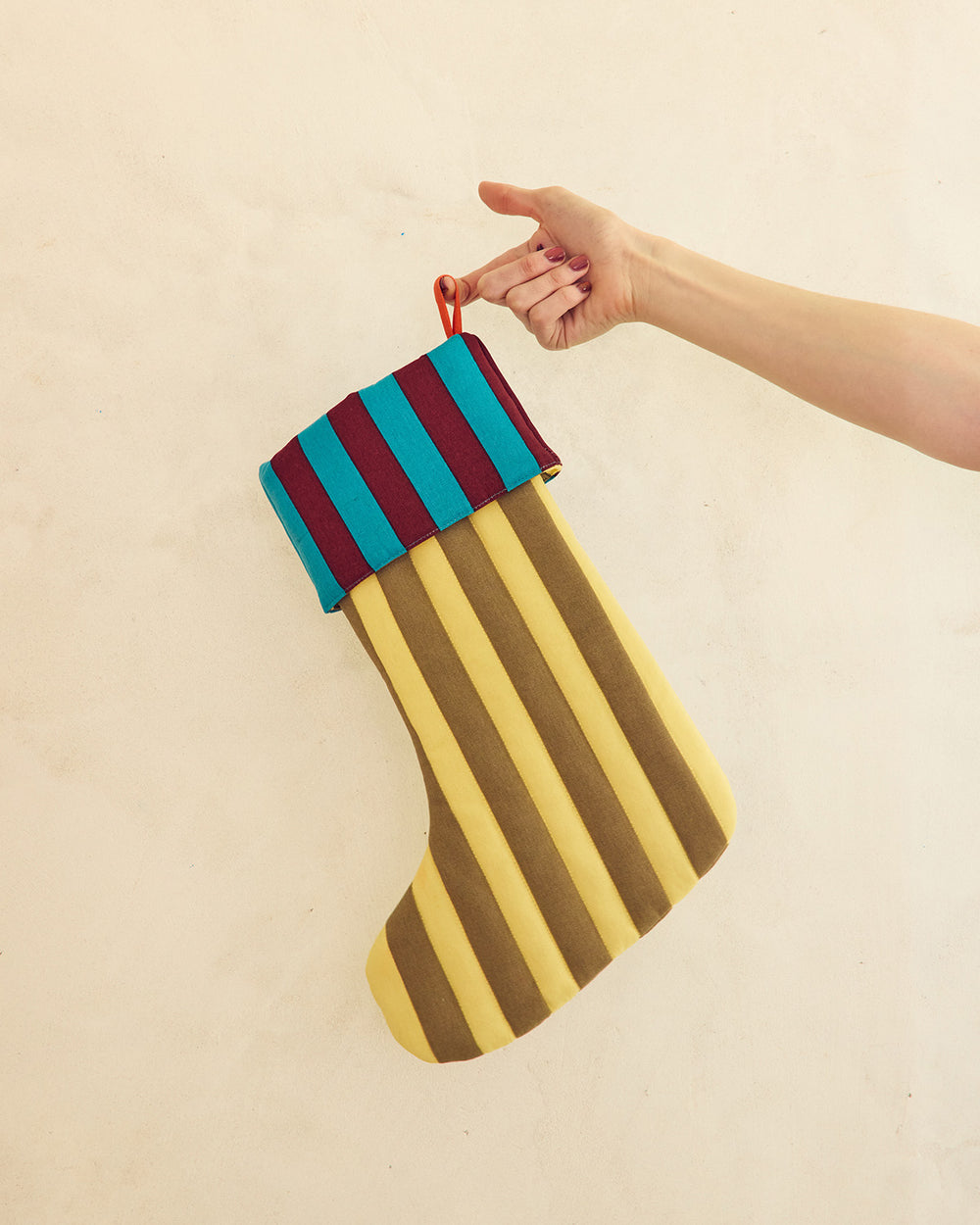 A hand holding a striped fabric Christmas stocking with alternating yellow and brown vertical stripes, a teal and burgundy striped cuff, and a red hanging loop, against a plain beige wall. 