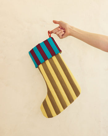 A hand holding a striped fabric Christmas stocking with alternating yellow and brown vertical stripes, a teal and burgundy striped cuff, and a red hanging loop, against a plain beige wall. 