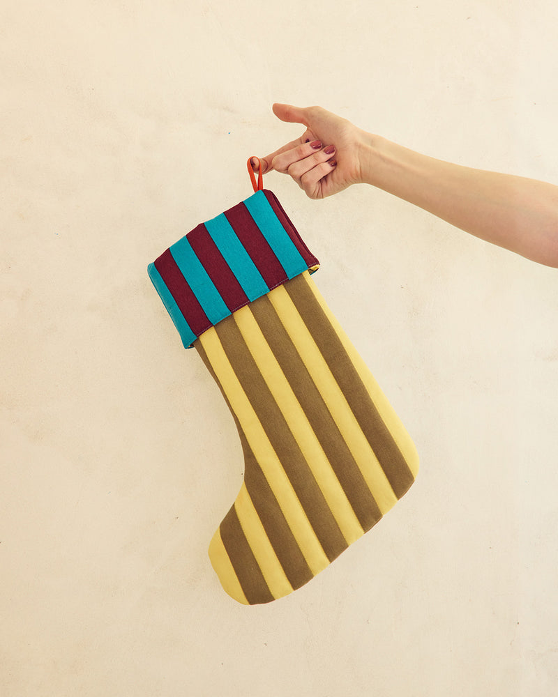 A hand holding a striped fabric Christmas stocking with alternating yellow and brown vertical stripes, a teal and burgundy striped cuff, and a red hanging loop, against a plain beige wall. 