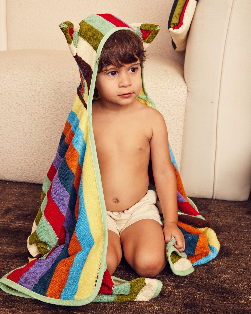 Child wearing a colorful striped towel with animal ears, sitting on the floor. 