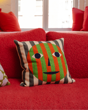["Dusen Dusen Everybody Face Pillow. Embroidered pillow in multi-colored Face print. Full embroidery on front and back. 100% cotton, bottom zip. on red couch", " Feeling lonely? Stare at this pillow!"]