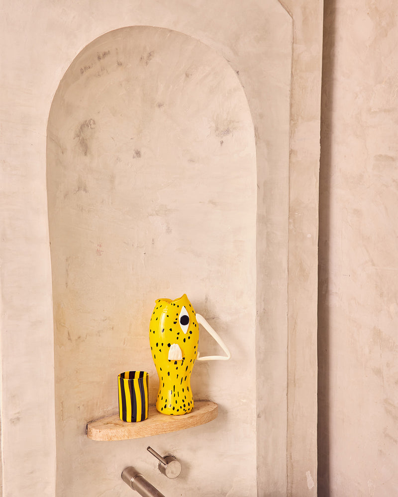 Yellow vase with black patterns on a wooden shelf against a beige wall.