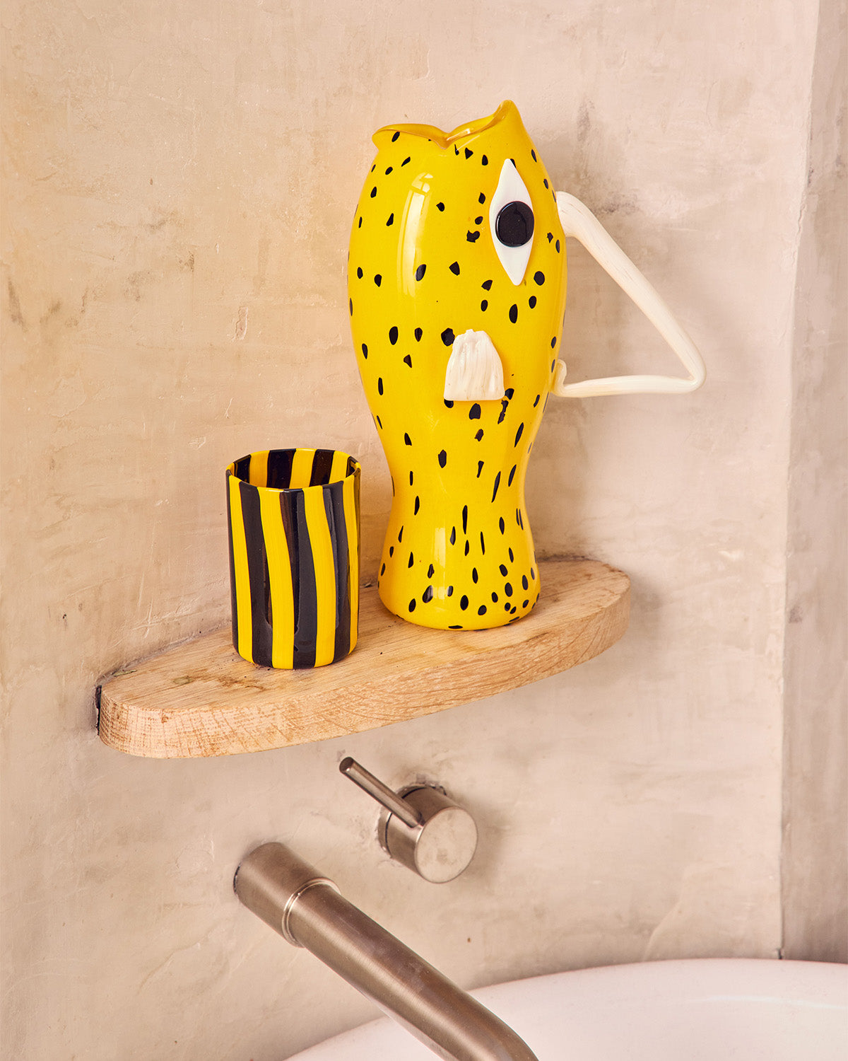 Yellow fish-shaped glass Pitcher and black and yellow striped cup on a wooden shelf in a plaster niche 