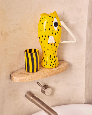 Yellow fish-shaped glass Pitcher and black and yellow striped cup on a wooden shelf in a plaster niche 