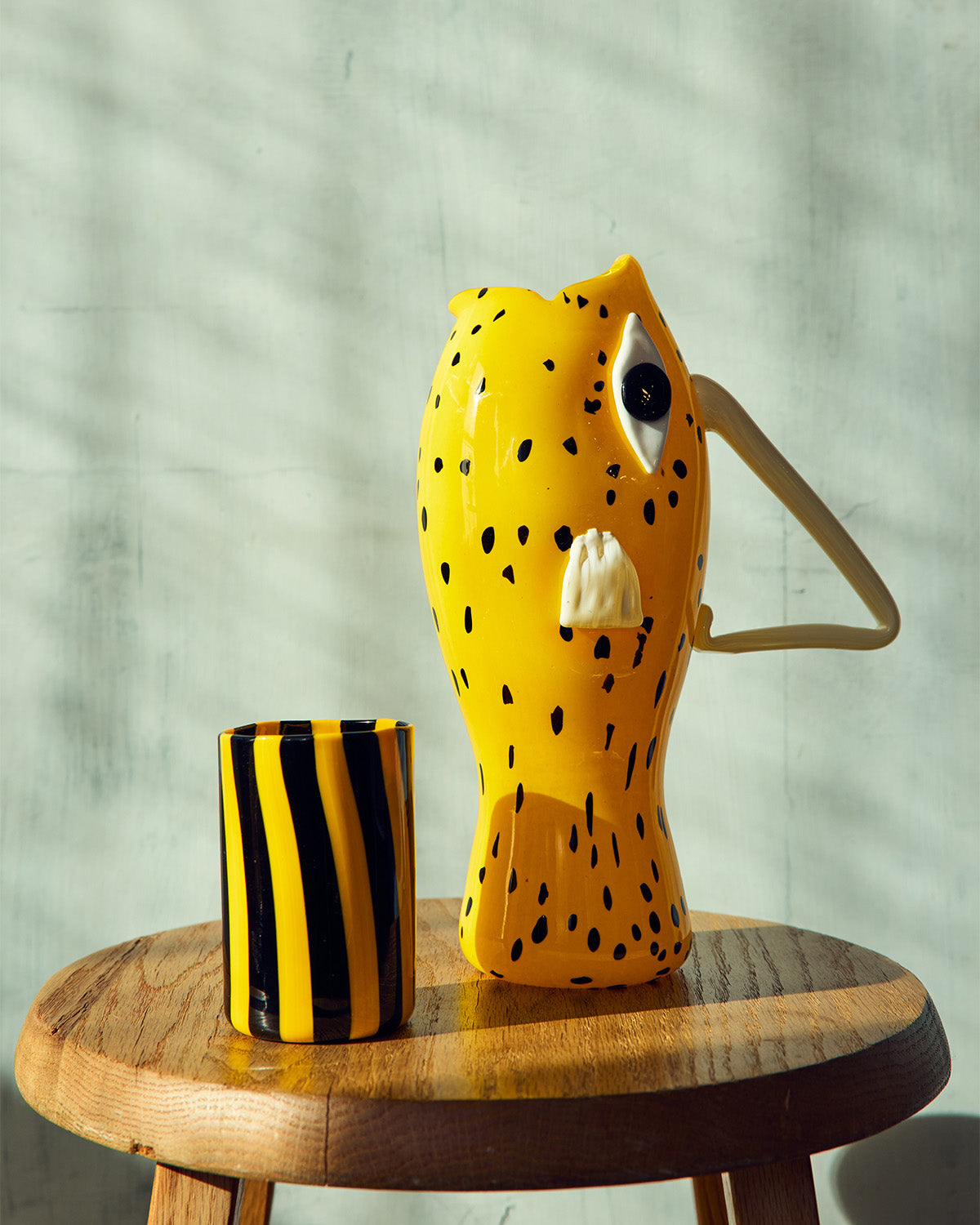 Yellow murano glass vase with black spots and a black and yellow striped cup on a wooden stool against a light green wall .
