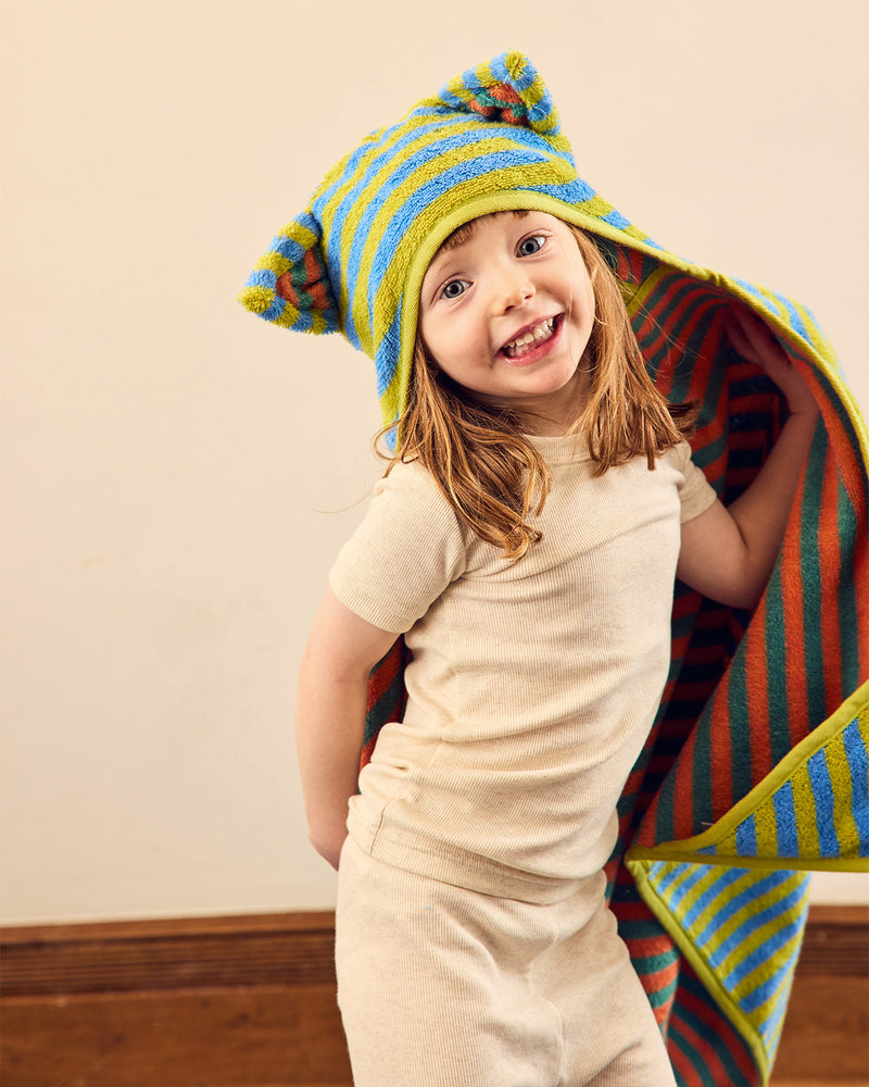 A young child wearing a cream-colored short-sleeve t-shirt and matching pants, smiling while wrapped in a colorful striped bath towel with a hood shaped like cat ears. 