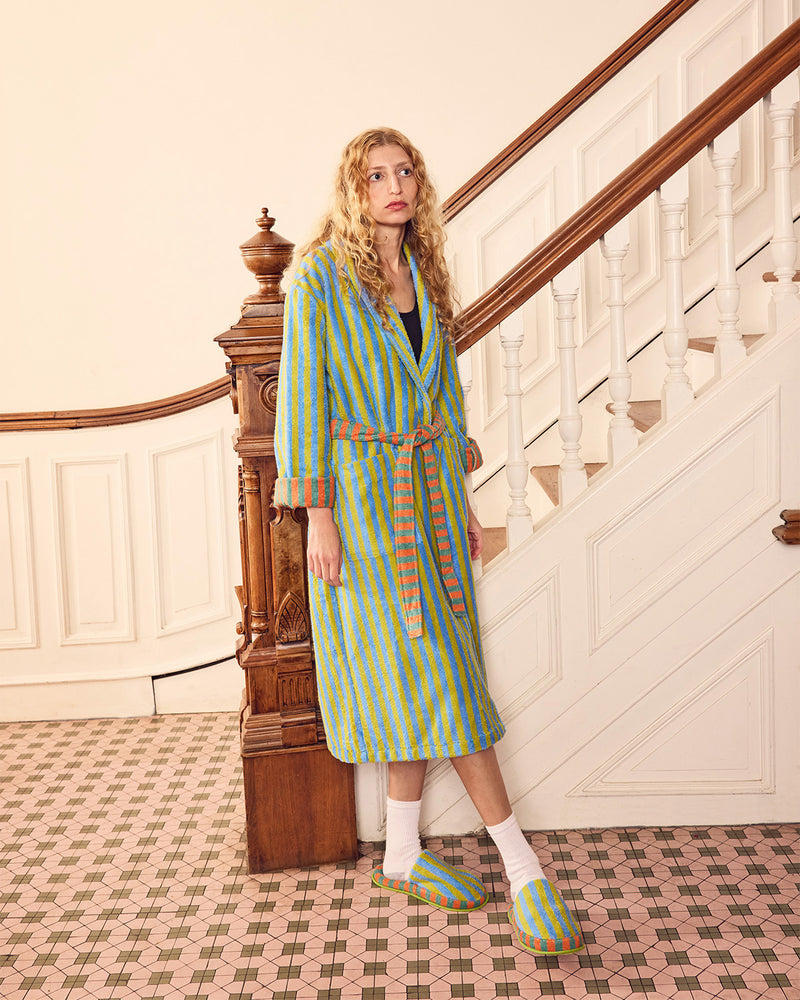 A woman stands on a patterned tile floor next to a wooden staircase, wearing a green and yellow striped bathrobe with a matching belt, white socks, and matching striped slippers.