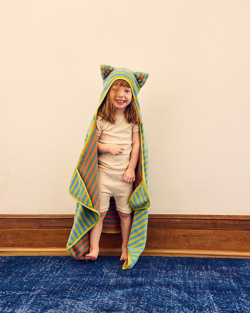 A young child stands barefoot indoors wearing a colorful striped hooded towel with ears, draped over their body. The towel features diagonal stripes in yellow, blue, orange, and green.