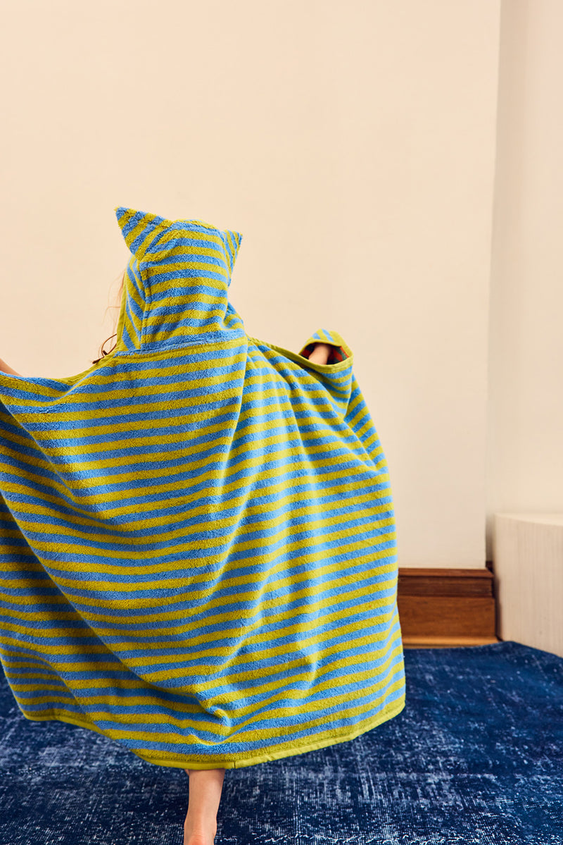A person wearing a hooded towel poncho with yellow and blue horizontal stripes, shown from behind in an indoor room with a cream wall and blue rug.