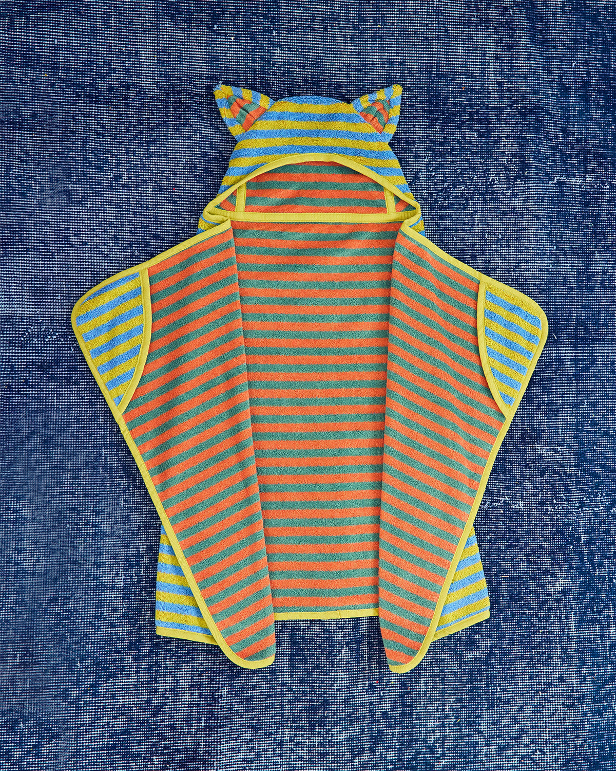 A children's hooded cape laid flat on a textured blue fabric background. The garment has bright orange and yellow horizontal stripes, a yellow hood with ear-like loops, and yellow trim along the edges.