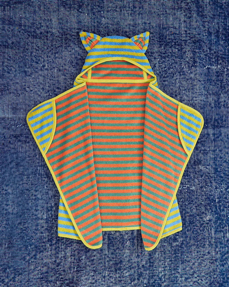 A children's hooded cape laid flat on a textured blue fabric background. The garment has bright orange and yellow horizontal stripes, a yellow hood with ear-like loops, and yellow trim along the edges.