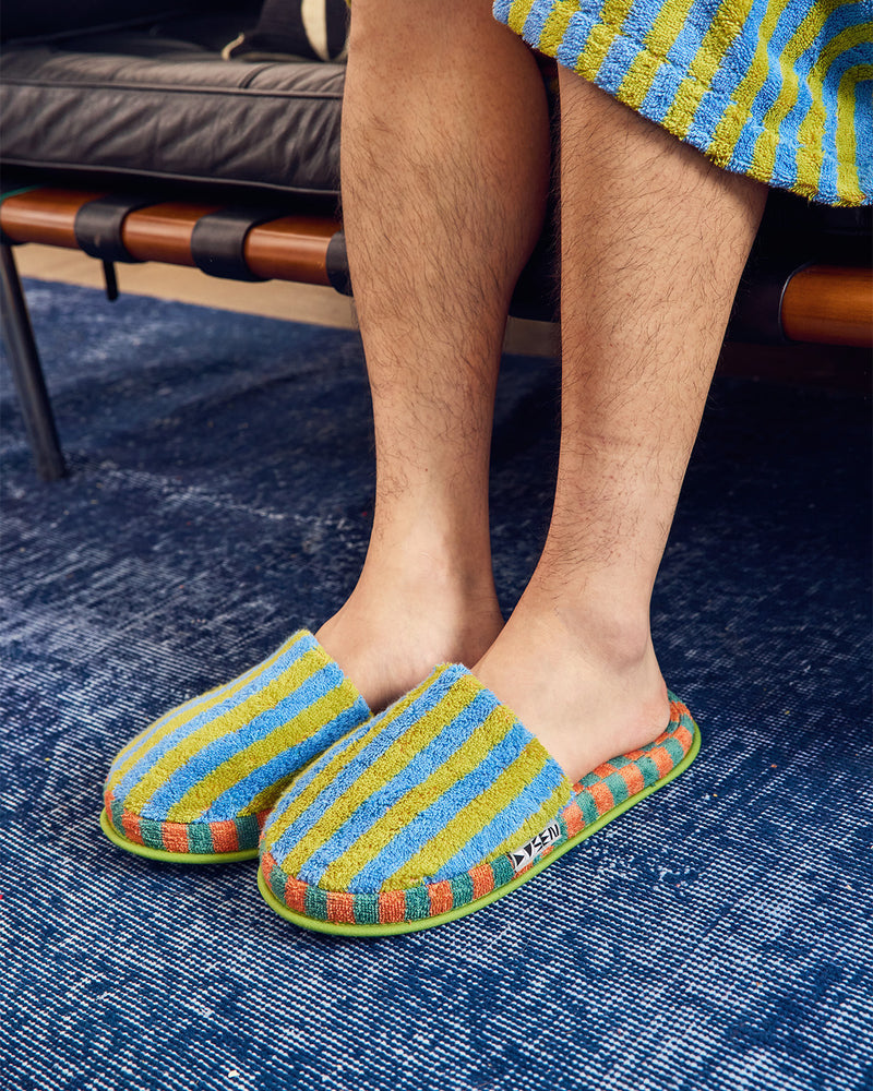 A person sitting on a bench wearing colorful striped terry cloth slip-on slippers with blue, yellow, orange, and green stripes, standing on a textured blue rug. 