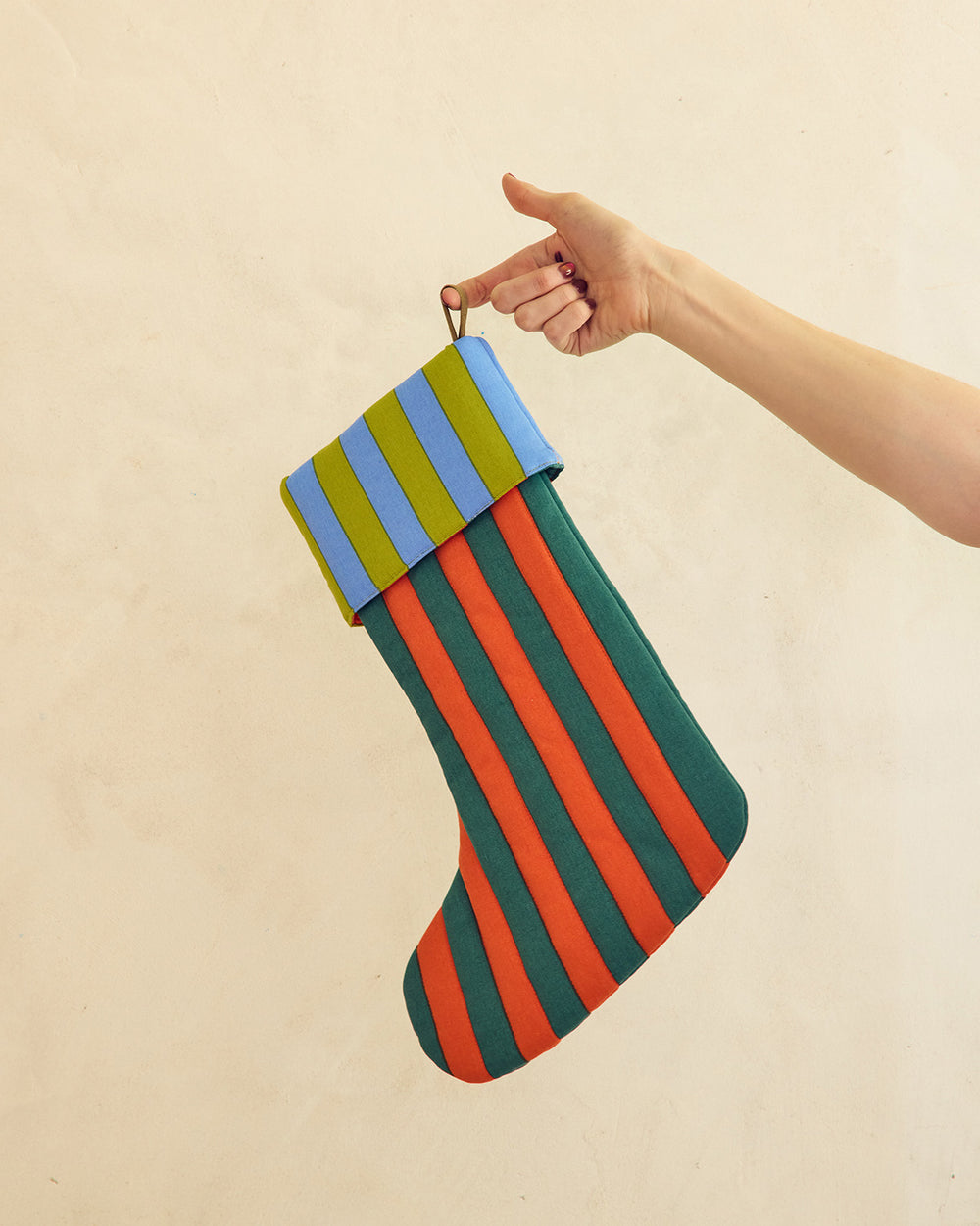 A hand holds a striped fabric Christmas stocking against a plain beige wall. The stocking has alternating vertical stripes of dark green, orange-red, and light blue, with a matching striped cuff and a small metal hanging loop. 