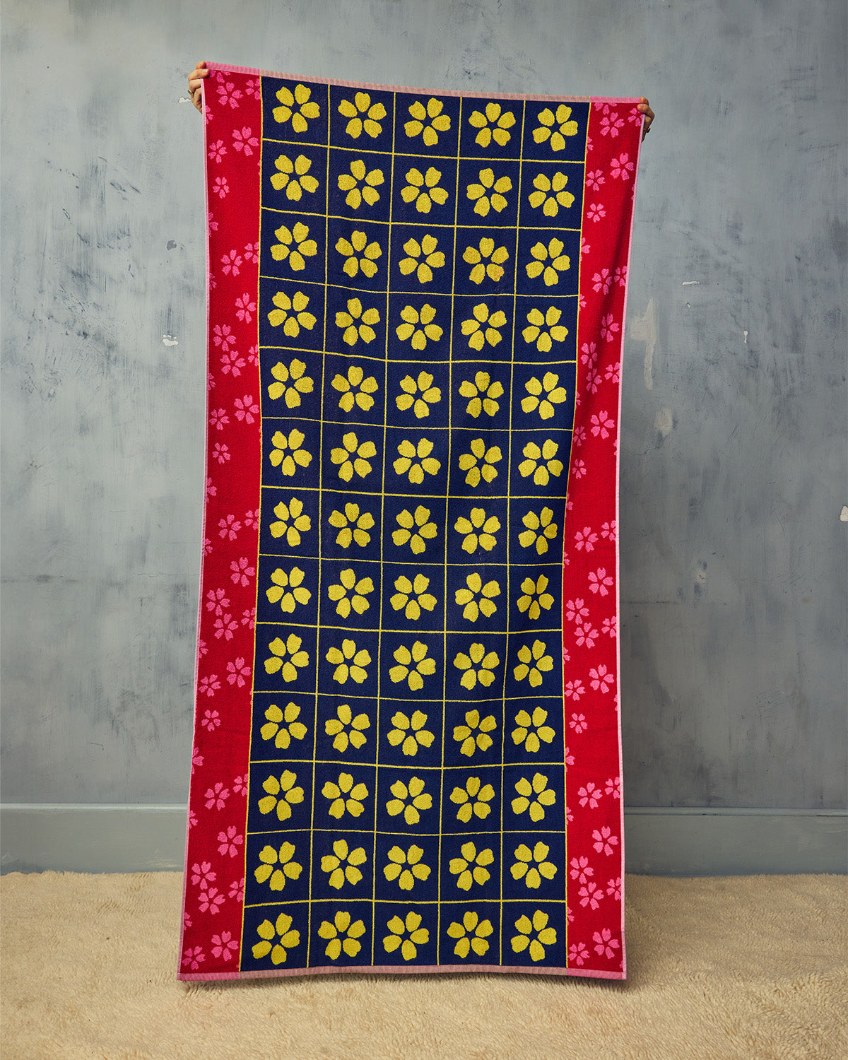 Towel with yellow floral pattern on a blue background, held against a gray wall.