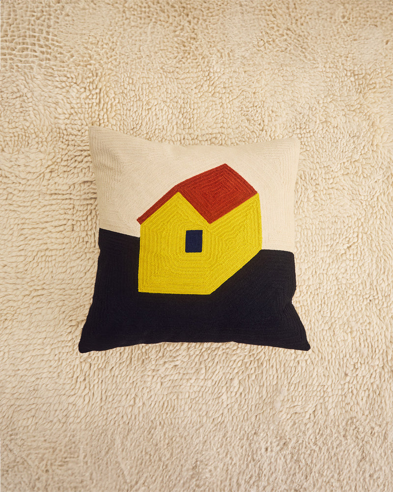 A square decorative throw pillow featuring a stylized house design with a red roof, yellow body, and black base, set against a textured cream background.