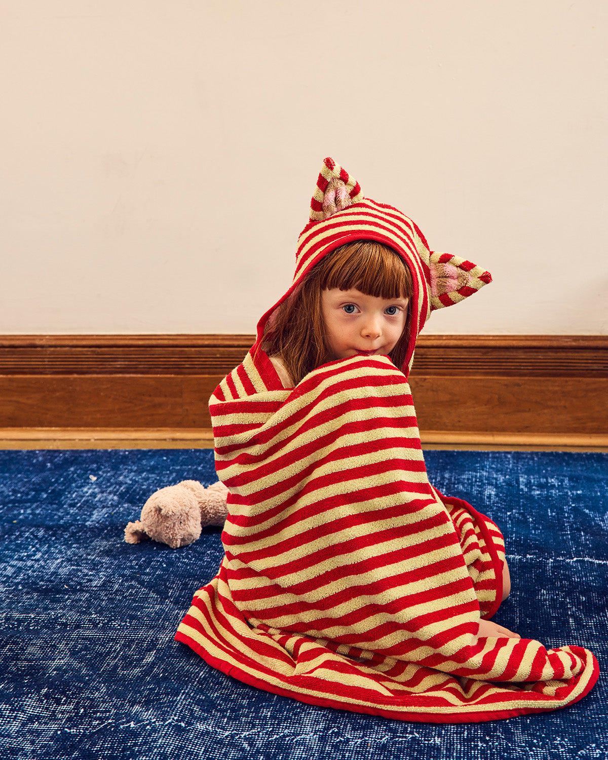 A child sitting on a blue rug wrapped in a red and cream striped hooded towel with cat-ear details, against a beige wall and wood baseboard. 
