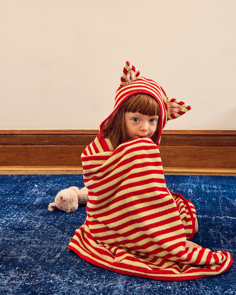 A child sitting on a blue rug wrapped in a red and cream striped hooded towel with cat-ear details, against a beige wall and wood baseboard. 