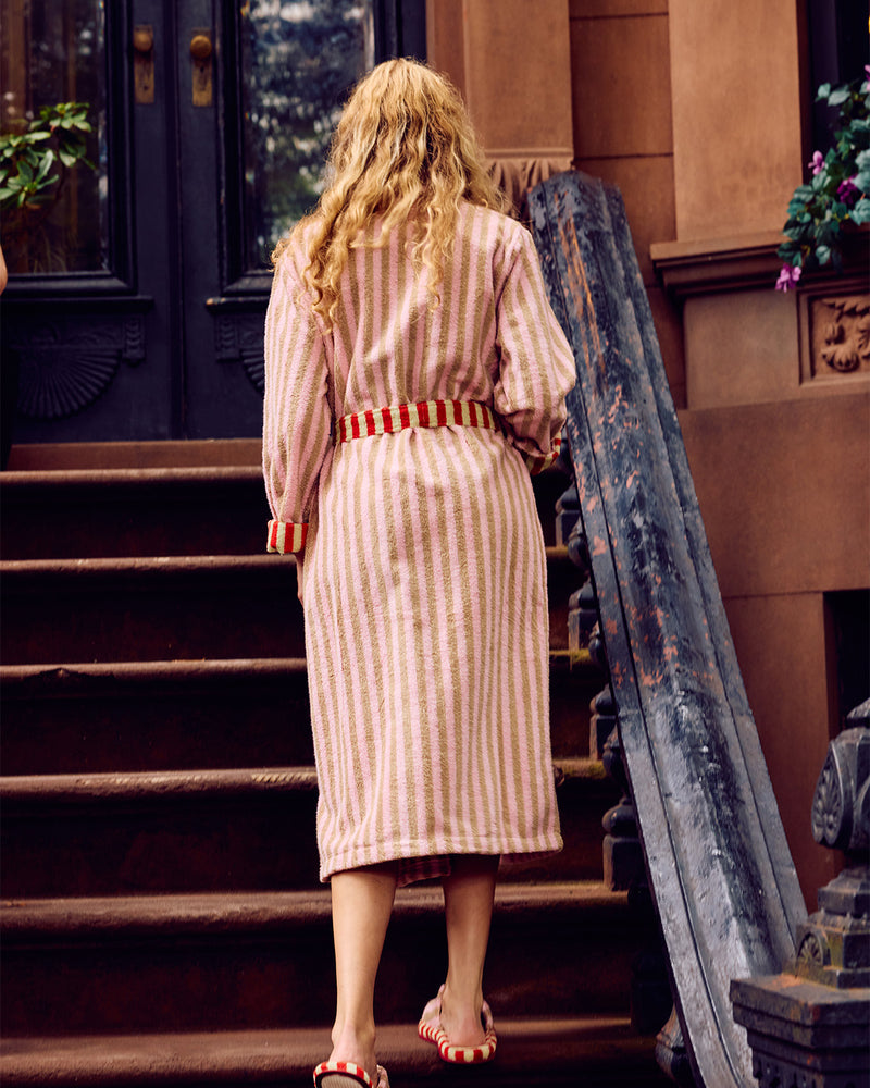 A woman viewed from behind wearing a pink and white vertically striped robe with a red and white striped belt, standing on outdoor stone steps in front of a brownstone building.