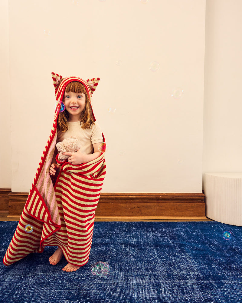 A young child smiling while wrapped in a red and white striped hooded blanket with ears, standing on a blue rug in an indoor room.