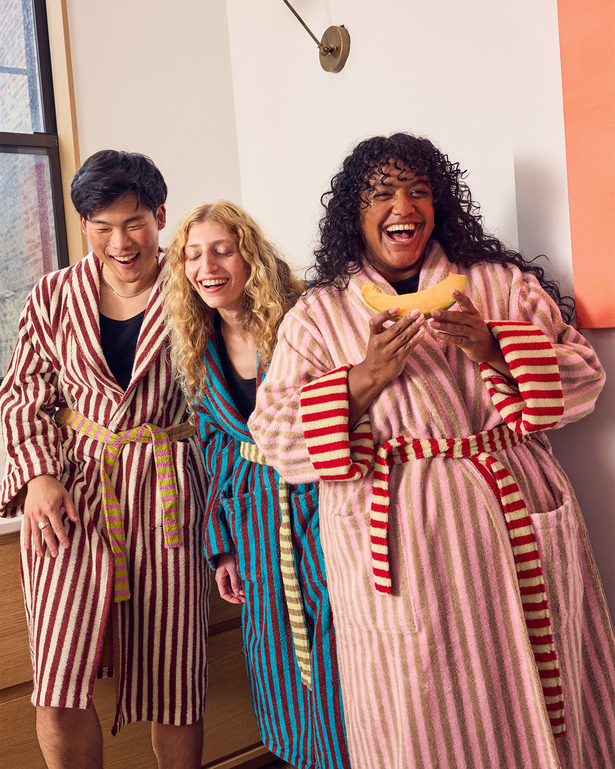 Three people laughing in matching striped bathrobes in a brightly lit indoor setting. The robe on the left is red and white vertically striped with a tan belt, the center robe is blue and white vertically striped with a gold belt, and the robe on the right is pink and white vertically striped with red stripes on the cuffs and a red-and-white striped belt.