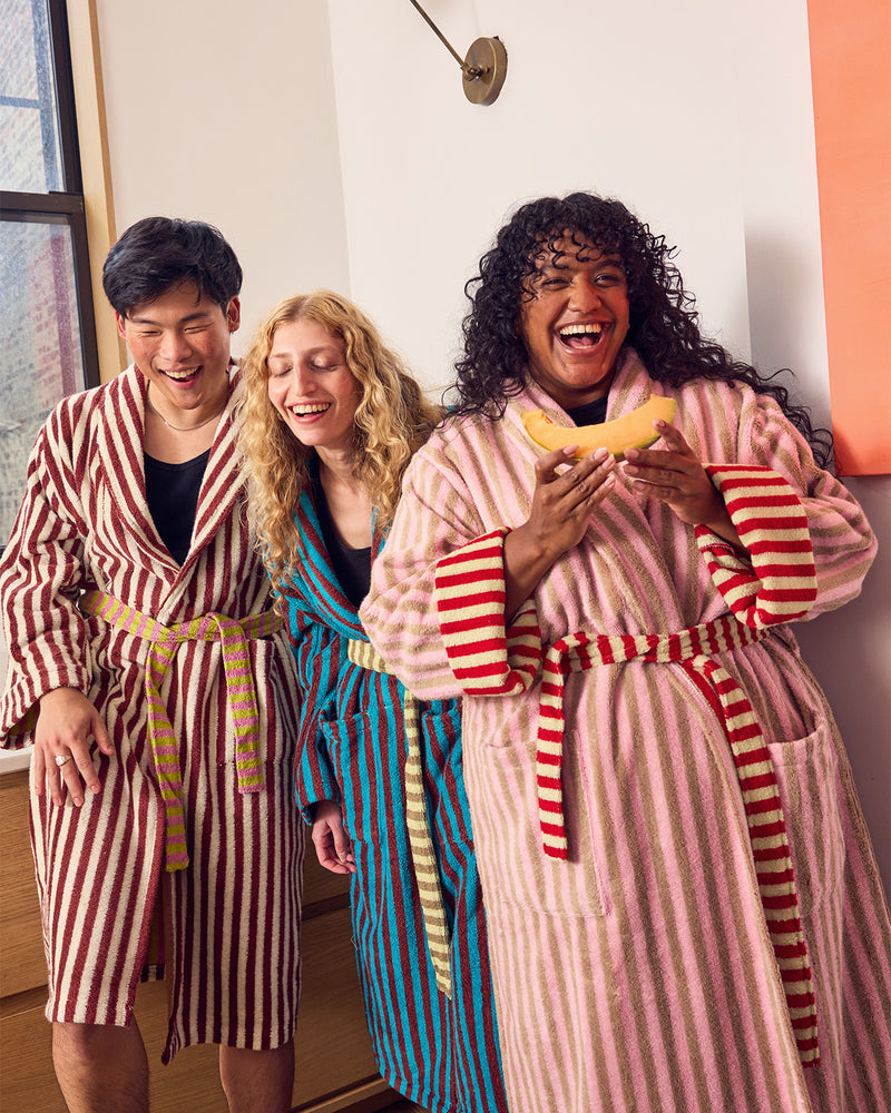 Three people laughing in matching striped bathrobes in a brightly lit indoor setting. The robe on the left is red and white vertically striped with a tan belt, the center robe is blue and white vertically striped with a gold belt, and the robe on the right is pink and white vertically striped with red stripes on the cuffs and a red-and-white striped belt.