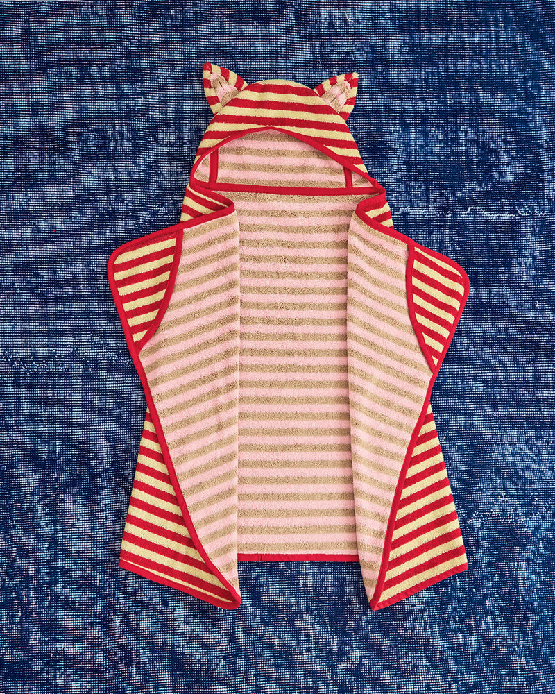 A baby hooded towel or wrap laid flat on a textured blue fabric background. The product has a pale pink and cream striped body, red trim, and ear details on the hood.