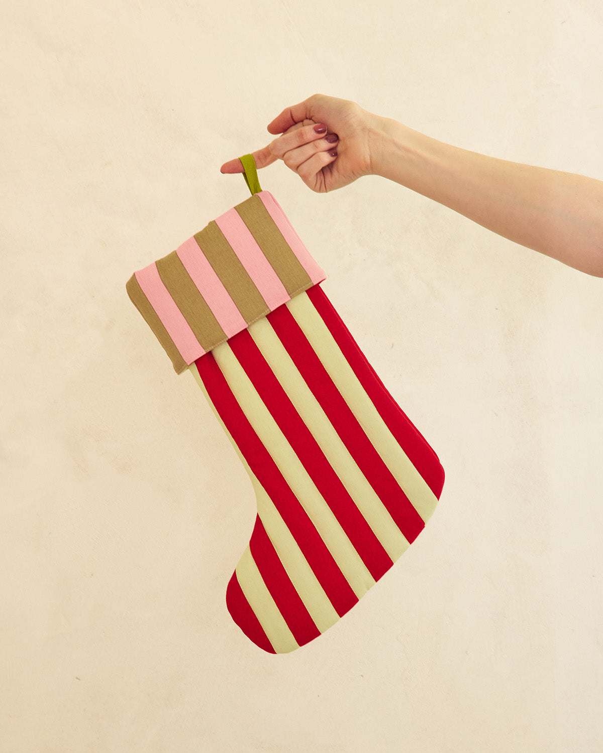 A striped fabric Christmas stocking held by a person's hand against a plain beige wall. The stocking has alternating red and cream stripes on the main body and pink and tan stripes on the cuff, with a small green hanging loop. 