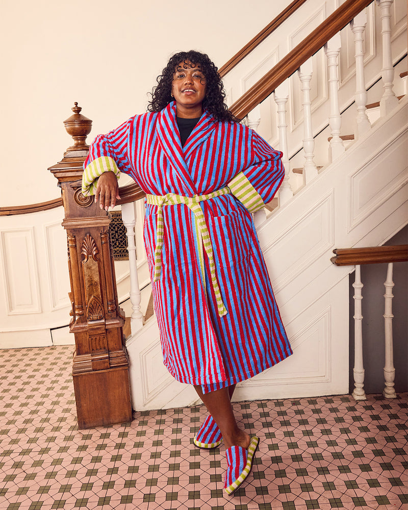 A woman stands in a grand staircase hall wearing a vibrant red, blue, and white striped bathrobe with yellow striped cuffs and a matching striped belt, along with matching striped slippers.