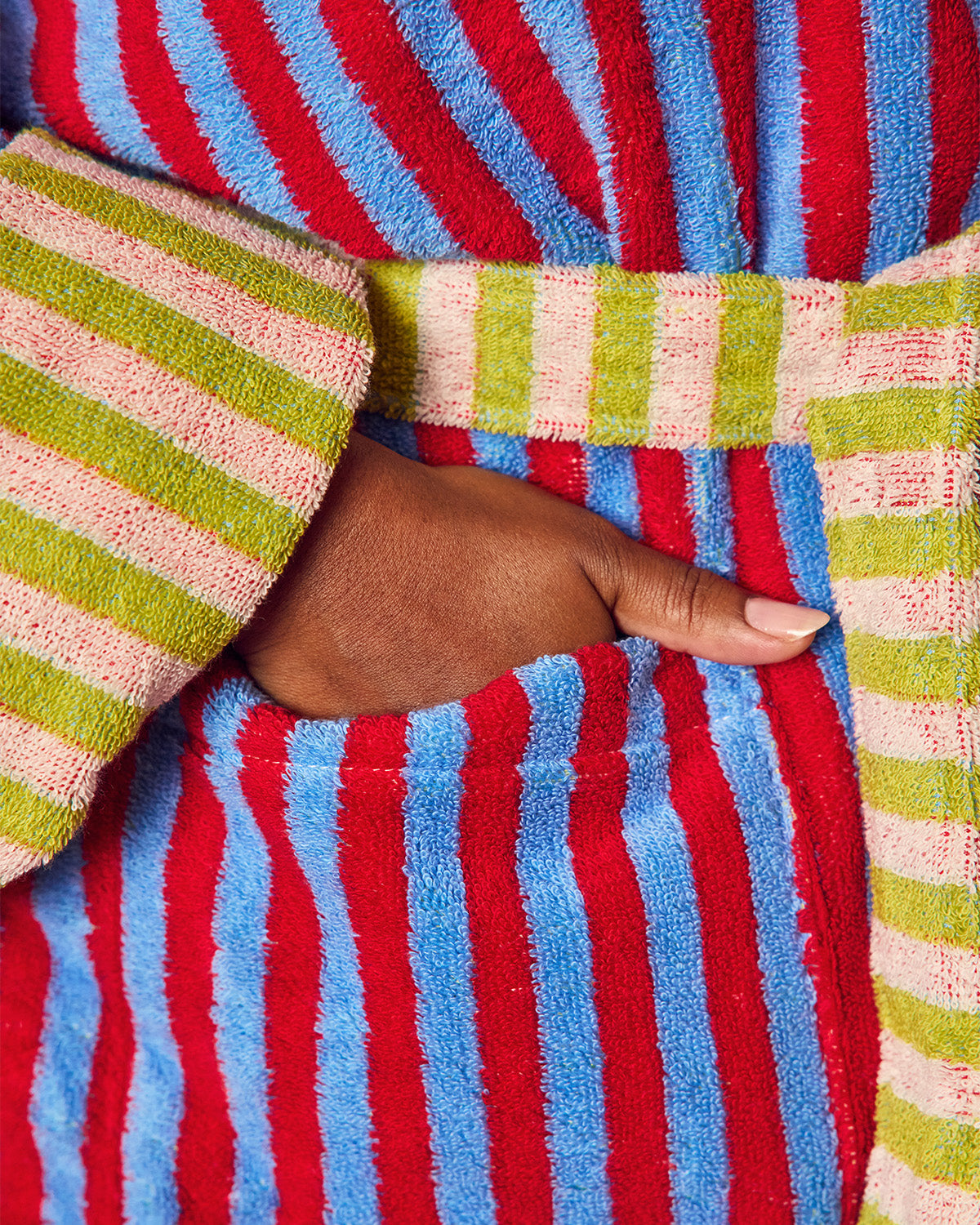 Close-up of a striped terry cloth robe with a hand tucked into a front pocket. The robe features vertical stripes in red, light blue, and lime green, with a matching striped belt at the waist.