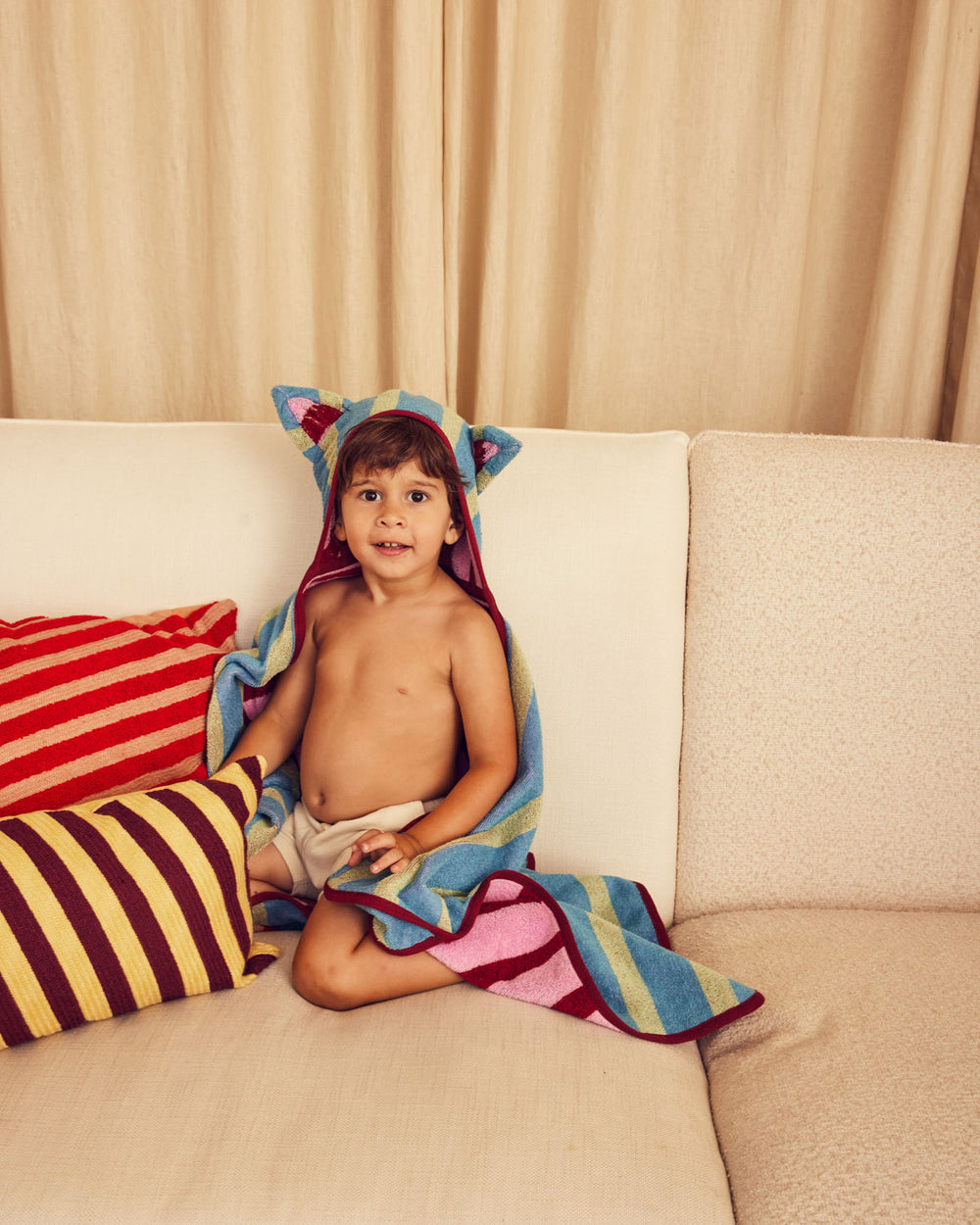Child sitting on a couch with a colorful towel draped over them, surrounded by striped pillows. 