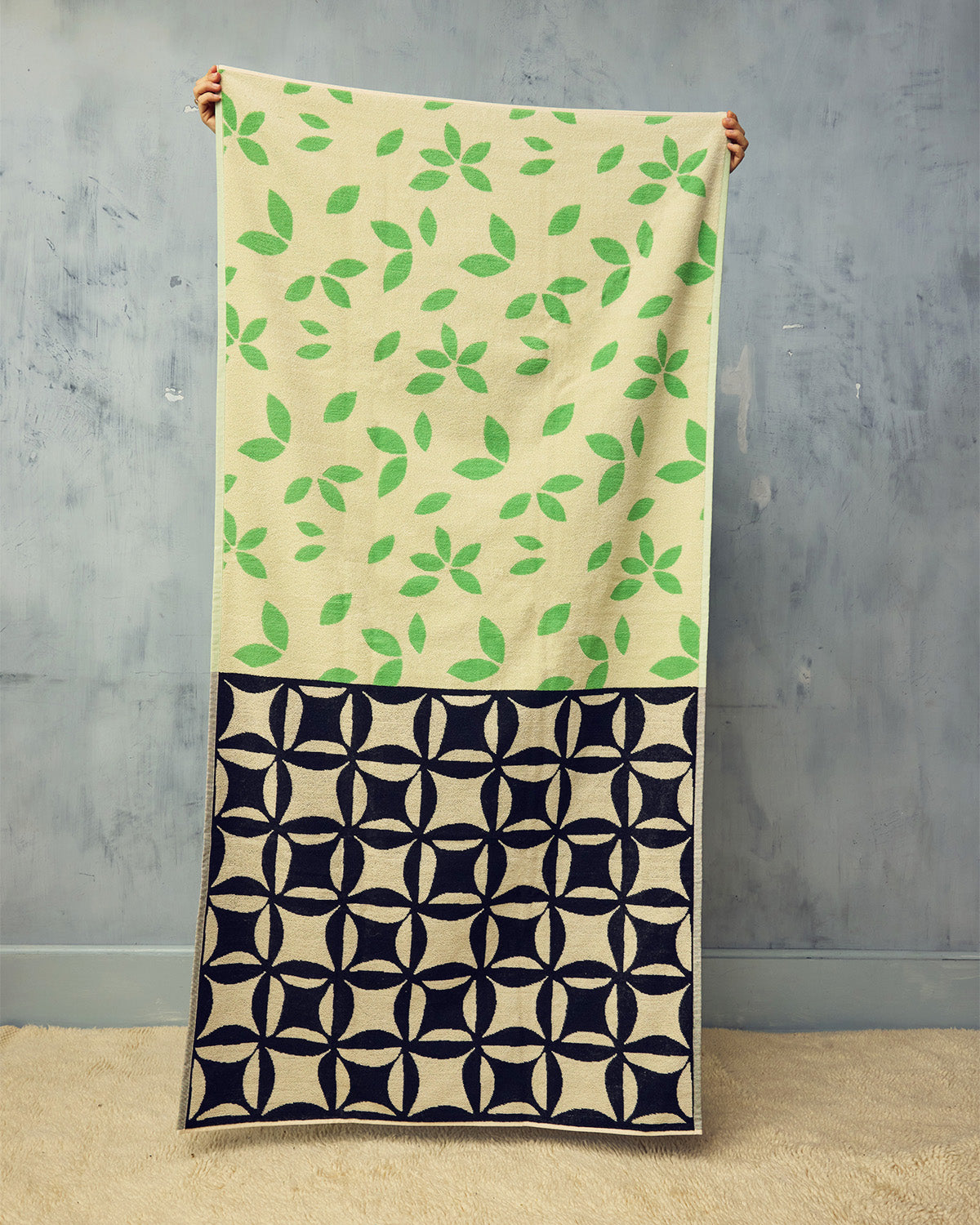Beige beach towel with green leaf pattern and black geometric pattern at the bottom, held against a gray wall.