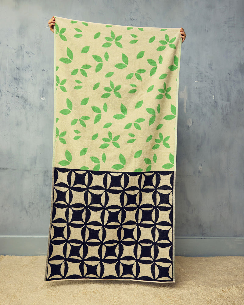 Beige beach towel with green leaf pattern and black geometric pattern at the bottom, held against a gray wall.