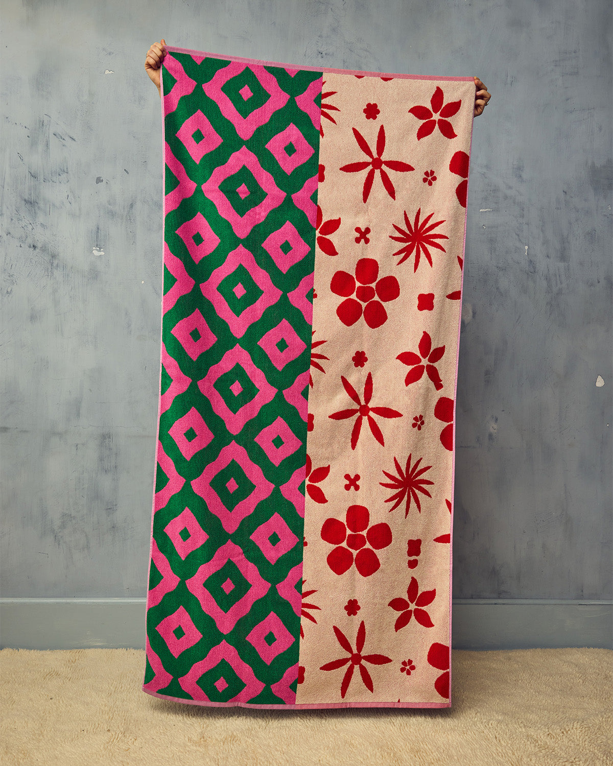 Peony towel in Ripple pattern and floral pattern being held up on opposite side in front of plaster wall