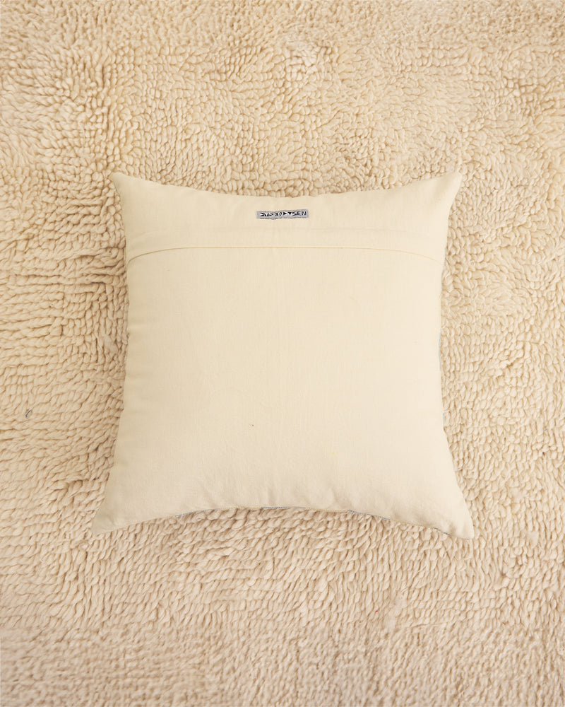 A square cream-colored throw pillow with a horizontal stitched seam across the middle, resting on a textured beige shag rug. 