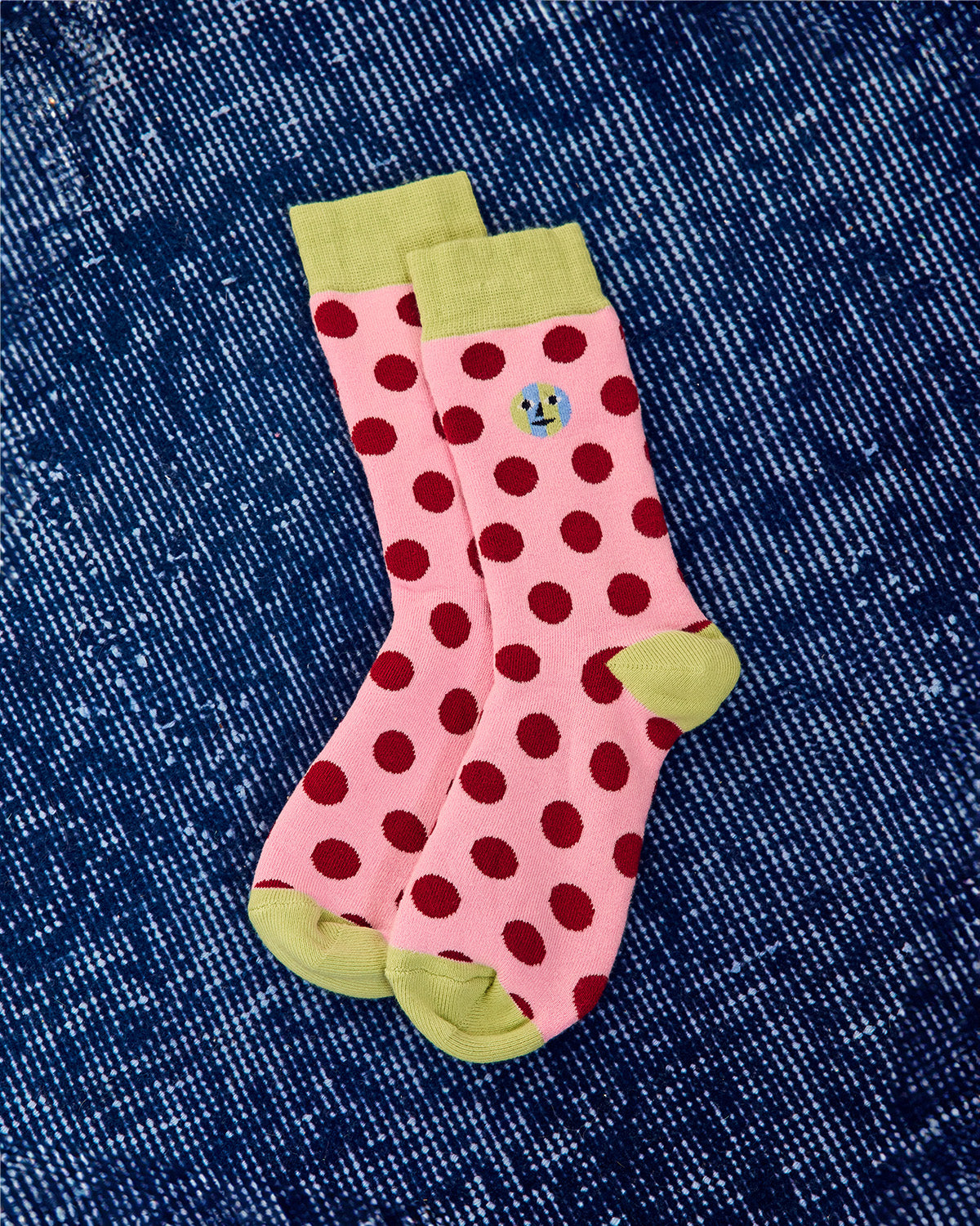 A pair of pink crew socks with dark red polka dots and light green heels, toes, and cuffs, laid flat on a blue denim background. 