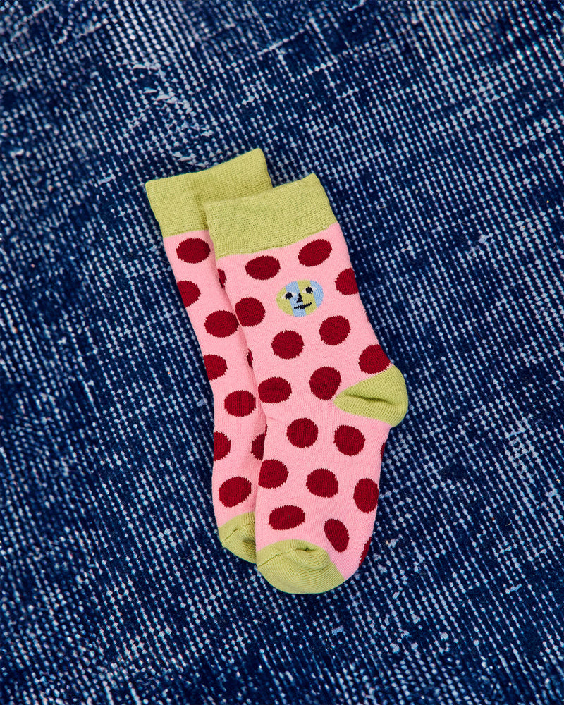 A pair of pink ankle socks with dark red polka dots, light green cuffs and toes, and a small cartoon face graphic on the side, laid flat on a blue denim background.