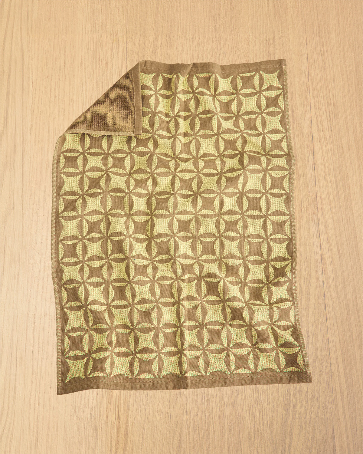 A rectangular woven textile with a repeating geometric medallion pattern in olive green and brown, laid flat on a light wood surface with one corner folded over.
