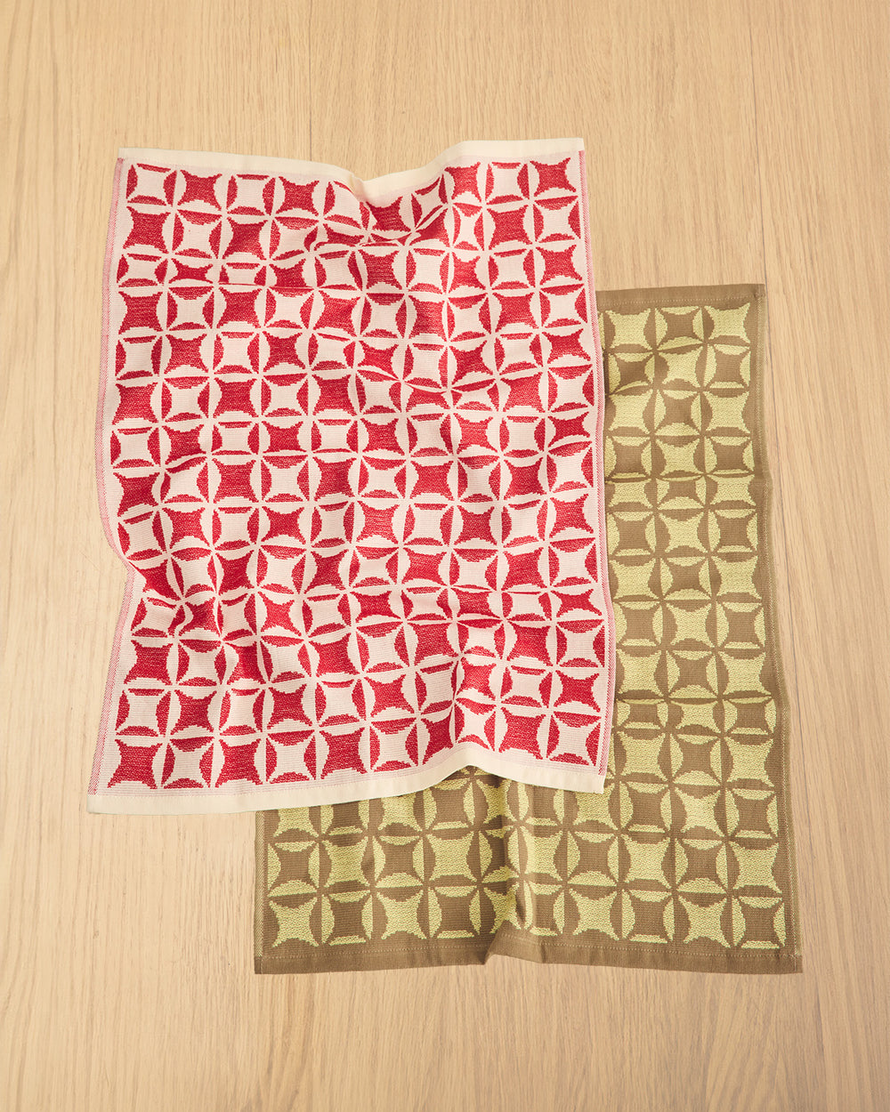 Two patterned square fabric pieces with geometric medallion motifs, one in red and off-white, and the other in olive green and brown, laid flat on a light wood surface. 