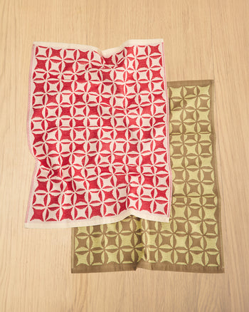 Two patterned square fabric pieces with geometric medallion motifs, one in red and off-white, and the other in olive green and brown, laid flat on a light wood surface. 