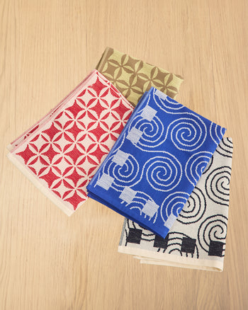 Four folded patterned fabric napkins or small table mats arranged on a light wood surface. The top visible item is tan with a repeating geometric motif, the center item is bright blue with white spiral and square patterns, and the bottom item is white with black spiral and black square patterns. 